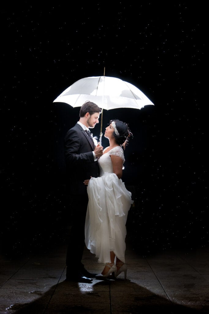 Les mariés sous un parapluie la nuit, avec les gouttes de pluie éclairées autour d’eux